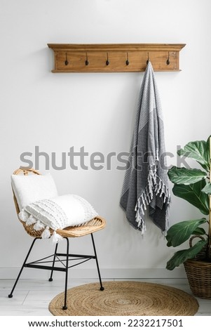 Boho interior with wicker rattan chair and cushion. Vertical shot of stylish vintage armchair in living room close to house plant ficus, home decoration and plaid on clothes hanger on white wall