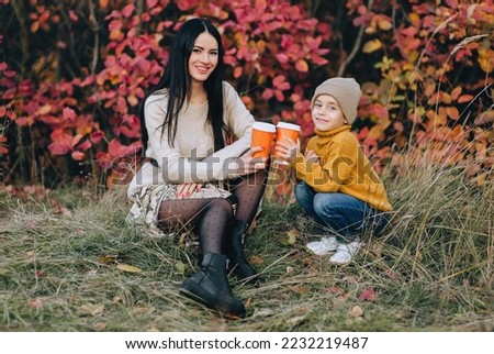 A girl, a woman and her little son are sitting on the green grass in autumn against the background of bushes, trees with red leaves with a cup of tea in their hands. Photo, smiling family.
