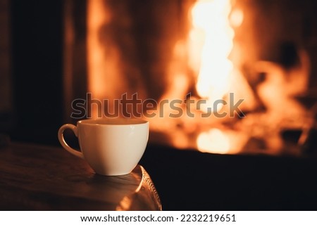 Hot chocolate in a mug on wooden table with cozy fireplace flame on the background. Winter season.