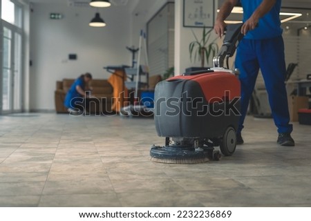 Cleaner cleans hard floor with scrubber machine while other cleaner cleans in the background Royalty-Free Stock Photo #2232236869