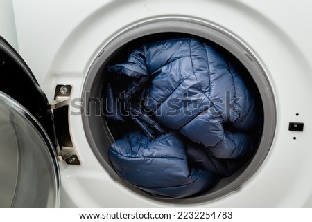 Blue winter jacket in the drum of open washing machine in laundry room. Washing dirty down jacket in the washer Royalty-Free Stock Photo #2232254783