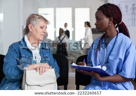 African american female doctor writing medication prescription for elderly sick woman in hospital lobby. Senior lady consulting general practitioner in geriatric clinic waiting area.