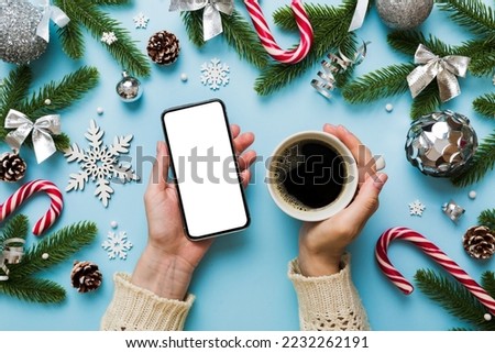 Above view of female hand holding smart phone with hot cup of coffee. Christmas decoration on colored background.