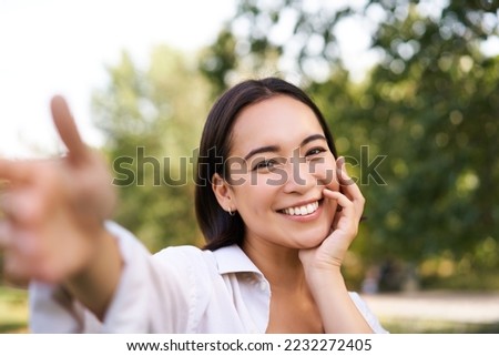 Smiling asian girl takes selfie, video chats, holds smartphone and speaks at camera, posing in park.