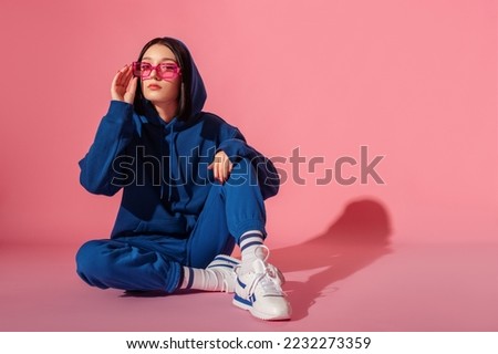 Young confident brunette woman wearing trendy blue suit with hoodie, trousers, pink color sunglasses, stylish white sneakers. Studio fashion portrait. Copy, empty space for text Royalty-Free Stock Photo #2232273359