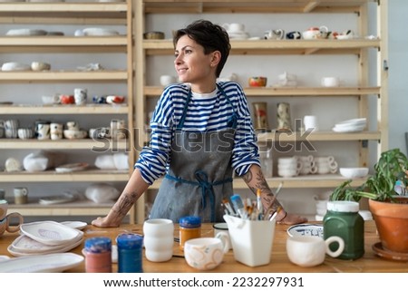Calm young female owner in pottery workshop looking at window and smiling, plans projects. Small business concept, ceramics studio. Successful italian woman entrepreneur with tattoos at workplace. Royalty-Free Stock Photo #2232297931