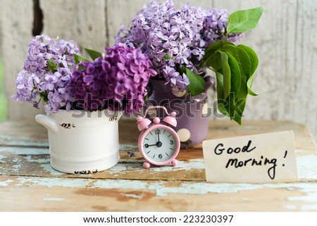 Two tone Lilac flowers in a ceramic pots white and purple, with pink vintage tiny alarm clock and a Good Morning note on a shabby wooden surface