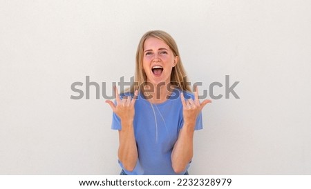 Emotive young woman with open mouth showing rock-n-roll gesture. Caucasian female model with blonde hair and blue eyes in blue T-shirt showing horns up. Lifestyle, rebellious concept