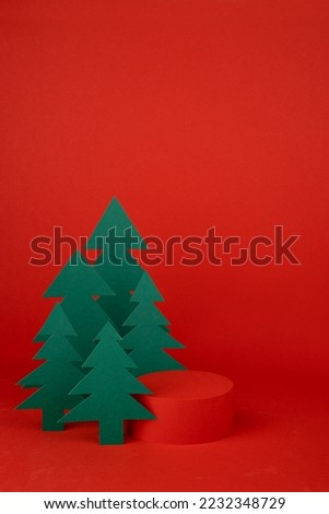 Christmas tradition background - red scene, cylinder podium mockup for presentation gifts, cosmetic products surrounded green paper spruce forest, vertical. New year template for card, poster, flyer.