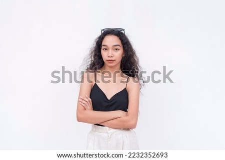 A slim and confident Filipino woman with long curly hair in her early 20s. Wearing a black spaghetti strap blouse and white pants. Arms crossed. Isolated on a white background.