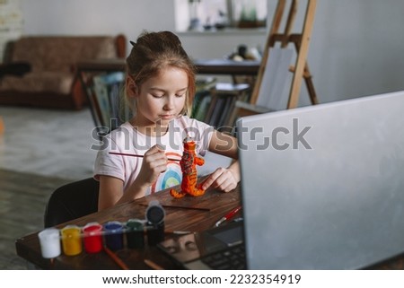 The beautiful cute girl creates crafts at home from the video lesson. A cheerful child is engaged in a hobby on a laptop. Elementary school student doing homework at homeschooling via video link. Royalty-Free Stock Photo #2232354919