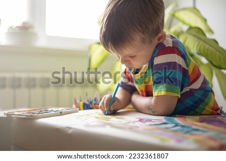 Confident cute male kid art drawing multicolored rainbow arch use paints and brush on table at kindergarten home interior. Focused baby boy enjoying artwork early development creative activity