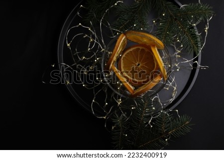 on a dark background and on a black tray stands a transparent glass with dry oranges and a branch of a Christmas tree and garlands on top of a little snow. for screensavers, postcards, labels, invitat