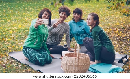 Pretty young women are taking selfie with smartphone during picnic in park sitting on grass, posing and laughing having fun. Nature, friendship and modern technology concept.