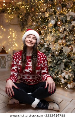 Beautiful girl in a Christmas sweater and a red Santa Claus hat sits near a luxurious Christmas tree. Smiling and looking at the camera. Celebration of new year, christmas