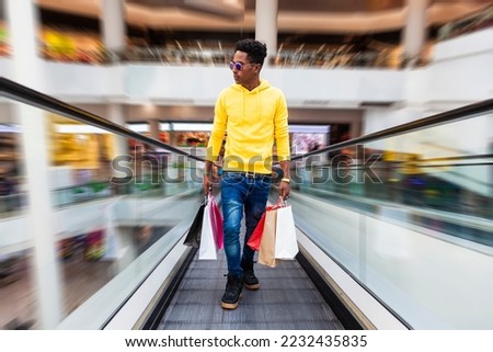 Portrait of a young African man dressed in a yellow sweatshirt, jeans and sunglasses holding shopping bags walking up a escalator ramp in a shopping mall. The background has motion effect.