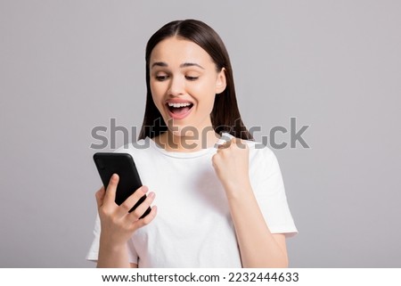 Inquisitive delighted young girl lady in white casual t-shirt standing on grey background holding cellphone smartphone in hands praying for favorite football team.