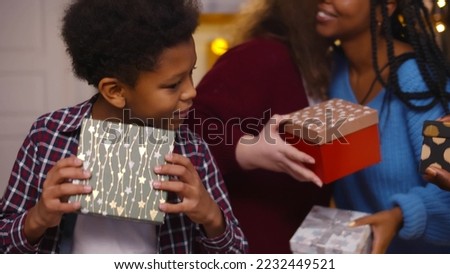 African-American boy opening christmas present box at party. Diverse friends exchanging and opening gifts celebrating new year together in decorated apartment