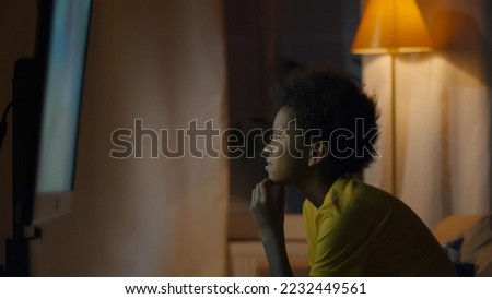 African-American kid sitting on sofa watching tv in living room. Side view of focused African-American preteen boy sitting in front of tv screen watching cartoon in evening at home