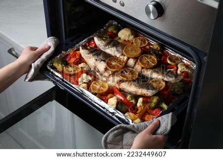 Woman taking out baking tray with sea bass fish and vegetables from oven, closeup Royalty-Free Stock Photo #2232449607