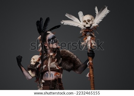 Shot of painted dark witch with fur and staff against grey background.