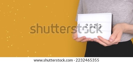 Banner with female hands hold lightbox with 2023 numbers in front of yellow background. New Year concept with place for your design.