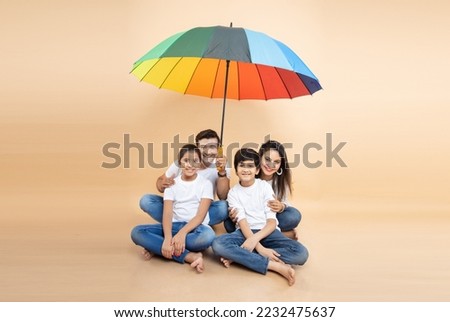 Happy indian family sitting under big  colorful umbrella isolated on beige background. parents and children together. hot summertime, Life and health insurance Safety, Holidays and vacations,  Royalty-Free Stock Photo #2232475637