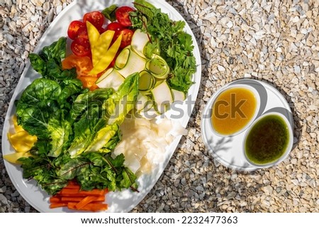 Vegetable plate with different types of greens and parmesan cheese. Nearby are two bowls with sauces. The dishes are on the gravel near the juniper pot.