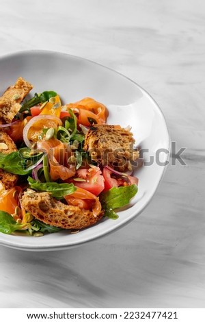 Summer salad with salmon, herbs, pumpkin seeds and slices of buckwheat bread in balsamic sauce. The salad lies in a light ceramic plate on a marble surface.