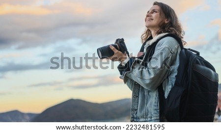 Adult Young cheerful woman traveler in a denim jacket with a backpack holds a camera in her hands while standing on the shore of mountain lake