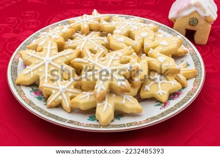 Plate with snowflake-shaped sugar icing Christmas cookies. 
Disposed on a red tablecloth with a house-shaped cookie as decoration.