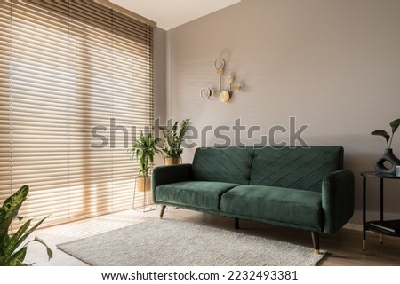 Elegant living room with big window wall behind wooden blinds, comfortable green sofa, carpet and decorative gold wall lamp Royalty-Free Stock Photo #2232493381