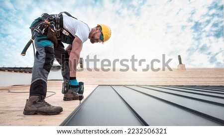A roofer with a safety harness and tool belt is working with a electric screwdriver on the roof. He is anchoring the metal roofing with a screws. Royalty-Free Stock Photo #2232506321
