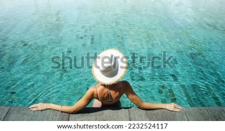 Back view of young woman wearing summer hat relaxing in big swimming pool with blue water on a sunny day. Copy space.
