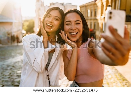 Two young beautiful smiling happy asian girls taking selfie on phone with raised hands near their cheeks , while standing on a sunny street