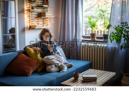 Young smiling cheerful woman in a warm sweater reading a book while sitting on the couch Royalty-Free Stock Photo #2232559683
