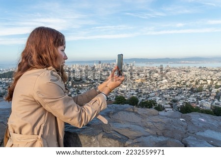 Asian tourist girl holding a smartphone to take a picture of the Twin Peaks viewpoint on a high mountain in the middle of San Francisco.