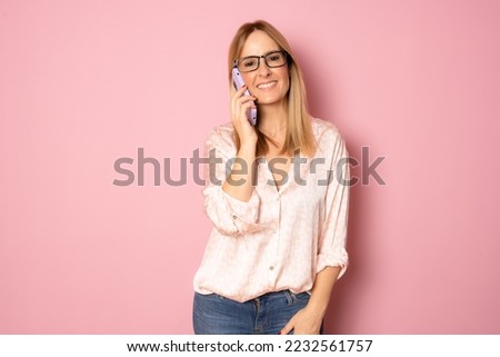 Picture of happy positive cheerful young woman talking on smartphone with excitement. Blonde woman in casual shirt using modern technologies. Isolated over pink background