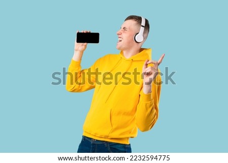 Portrait of overjoyed caucasian guy singing song and using cellphone as microphone, wearing wireless headset. Young man having fun over blue studio background