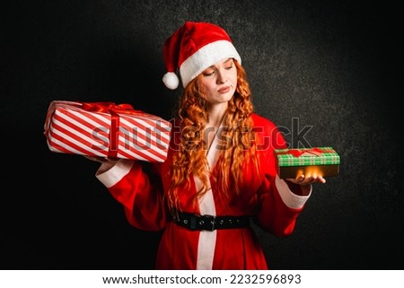 Portrait of a red-haired woman in a red hat and Santa Claus costume holding gift boxes on a dark black background.