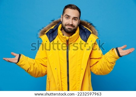 Young caucasian man 20s wear yellow down jacket spread hands shrugging shoulders looking puzzled, have no idea isolated on plain blue color background studio portrait. People winter lifestyle concept Royalty-Free Stock Photo #2232597963