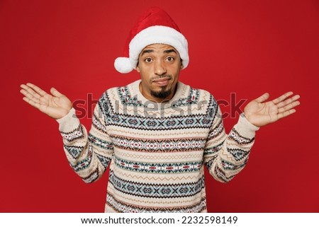 Merry young man wear Christmas sweater Santa hat posing spread hands shrugging shoulders looking puzzled, have no idea isolated on plain red background. Happy New Year 2023 celebration holiday concept