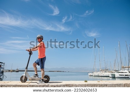 Elderly man in an orange jersey and protective helmet , rides on his electric scooter in front of the marina , isolated on sky background .