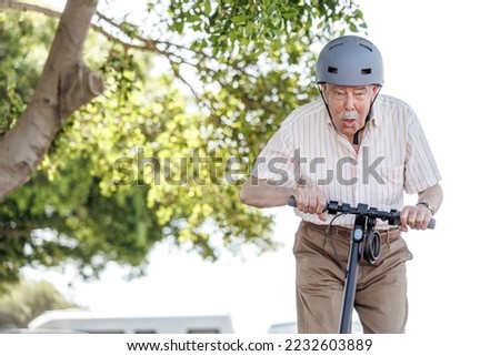 elderly man with helmet , electric scooter rides with busy expression in a bike path near a city park . Royalty-Free Stock Photo #2232603889