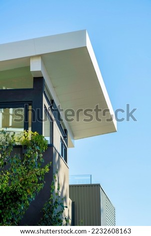 Decorative building facade with modern white roof extending over the house in late afternoon sun and front yard trees. In the neighborhood or in the city with blue house and sky background.