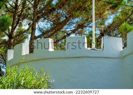 Top of building with castle style facade on turret or spire on structure with white paint and stucco exterior. In shade with touch of sun in late afternoon with tree and foliage and background plants.