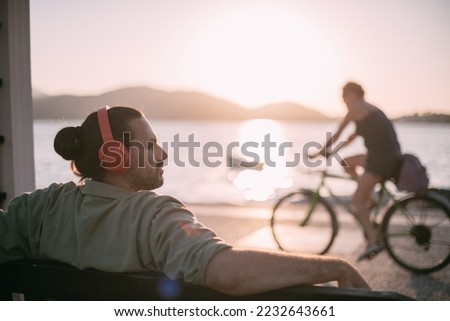 Portrait of a young man in bright large headphones listening to music, audiobook by the sea at sunset. A guy listens to a podcast, an audio tutorial sits relaxed on the ocean in the rays of the sun