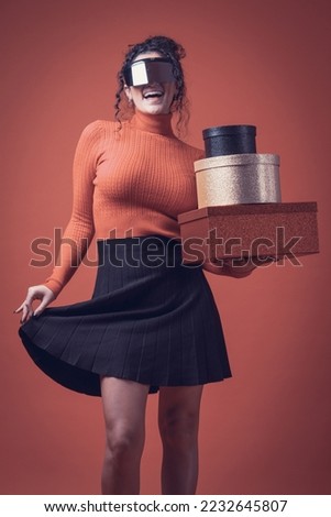 Excited girl smiles having fun with futuristic cyber glasses, glitter gold, black and red boxes, orange background, sweater and black skirt