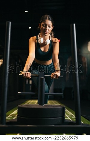 Young athletic Asian woman exercising with heavy training sled in a gym, gliding. Front view.