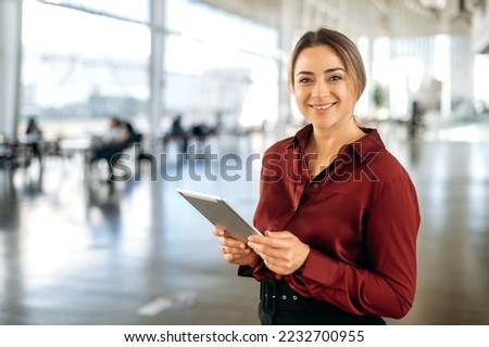 Portrait of positive beautiful intelligent caucasian business lady, recruiter, broker, company employee, in elegant clothes, holding a tablet in her hands, stand in the office, looks at camera, smiles Royalty-Free Stock Photo #2232700955